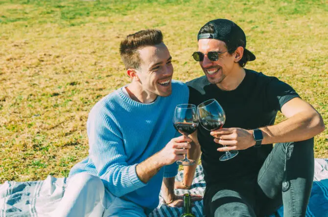 Pareja Gay Riendo Despreocupadamente Brindando con Copas de Vino Tinto en un Picnic en el Parque Sentados en la Hierba Verde